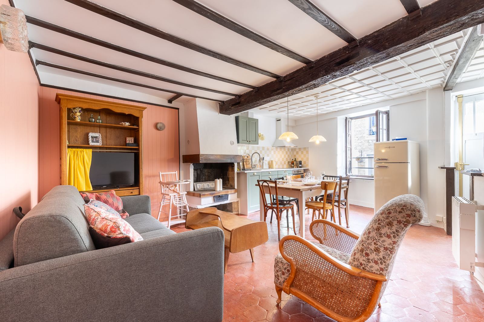 Le séjour : poutres anciennes, lambris rose, tomettes, fauteuil cannage fleuri, cheminée et cuisine vert sauge